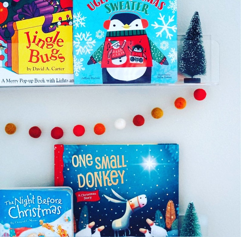 May include: A white wall with a garland of colorful felt balls. There are three bookshelves with children's books on them. The books are titled "Jingle Bugs", "Ugly Sweater", and "One Small Donkey". There are two small artificial Christmas trees on the shelves.