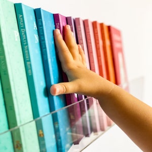 May include: A bookshelf filled with colorful paperback books. The books are arranged in a rainbow pattern, with the spines facing out. A hand reaches out from the right side of the image, touching the top of the books.