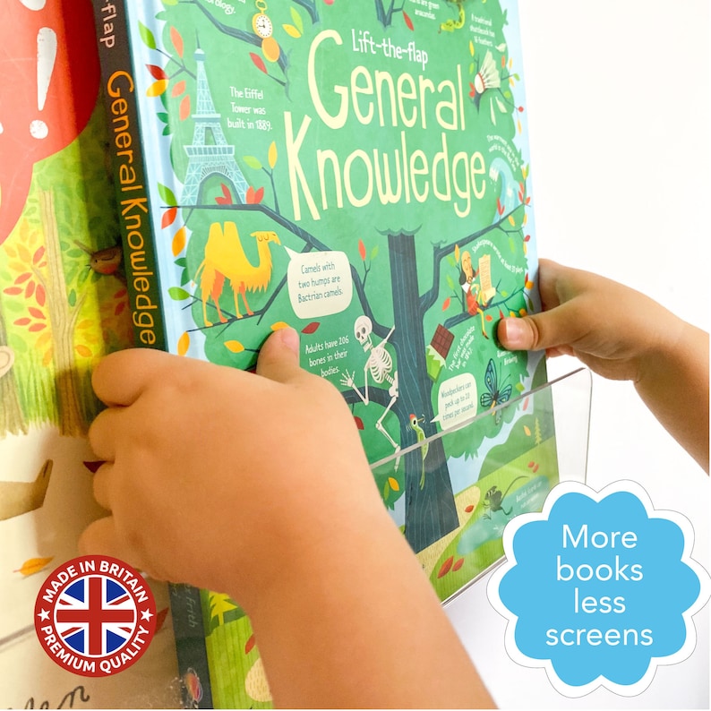 May include: A child's hand reaches for a lift-the-flap book titled "General Knowledge". The book features colorful illustrations of a camel, a skeleton, and a tree. The book is on a shelf with a sign that reads "More books less screens".