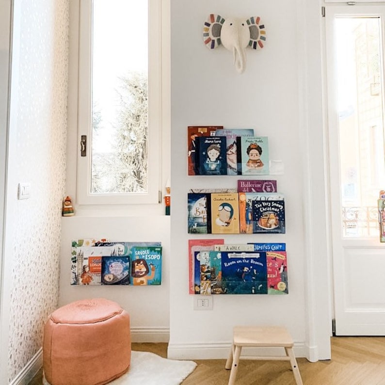 May include: A white wall with a bookshelf filled with colorful children's books. The bookshelf is mounted on the wall and has a white elephant decoration above it. There is a pink ottoman and a small wooden stool in front of the bookshelf.