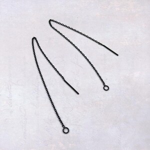 May include: A pair of black chain earrings. Each earring has a fine chain hanging from a small hoop at the bottom and a straight bar at the top. The earrings are set against a light background.