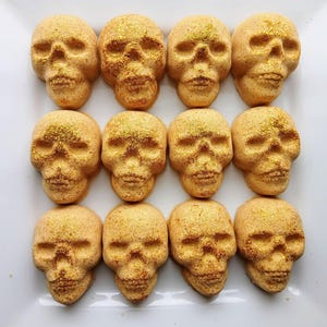 May include: Twelve skull-shaped treats arranged on a white surface. The treats are a light orange color with a textured surface and gold glitter accents. The skulls are evenly spaced, creating a symmetrical pattern.