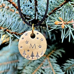 Puede incluir: Un collar con colgante redondo de color latón con un cordón negro. El colgante presenta la letra "M", estrellas y árboles. El collar cuelga frente a un fondo de agujas de pino azul-verdosas.