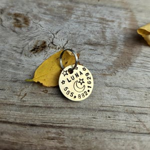 May include: A round brass pet identification tag featuring the stamped name LUNA, a crescent moon, and star icons, along with the phone number 565-882-1629. The metal disc includes a silver-toned split ring for attachment to a collar.