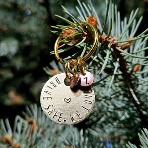 May include: A gold-colored keychain with a round pendant that reads "DRIVE SAFE. WE LOVE" around a heart symbol. It includes two smaller charms with the letters "Z" and "J". The keychain is hanging in front of a blurred green background.