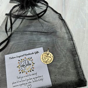 May include: A round gold-toned metal pet tag featuring the name SASHA, a crescent moon, and stars, with the phone number 555-174-2271 engraved. The tag is placed on a black sheer organza drawstring bag next to a white card with the text ReeJollie Gifts.