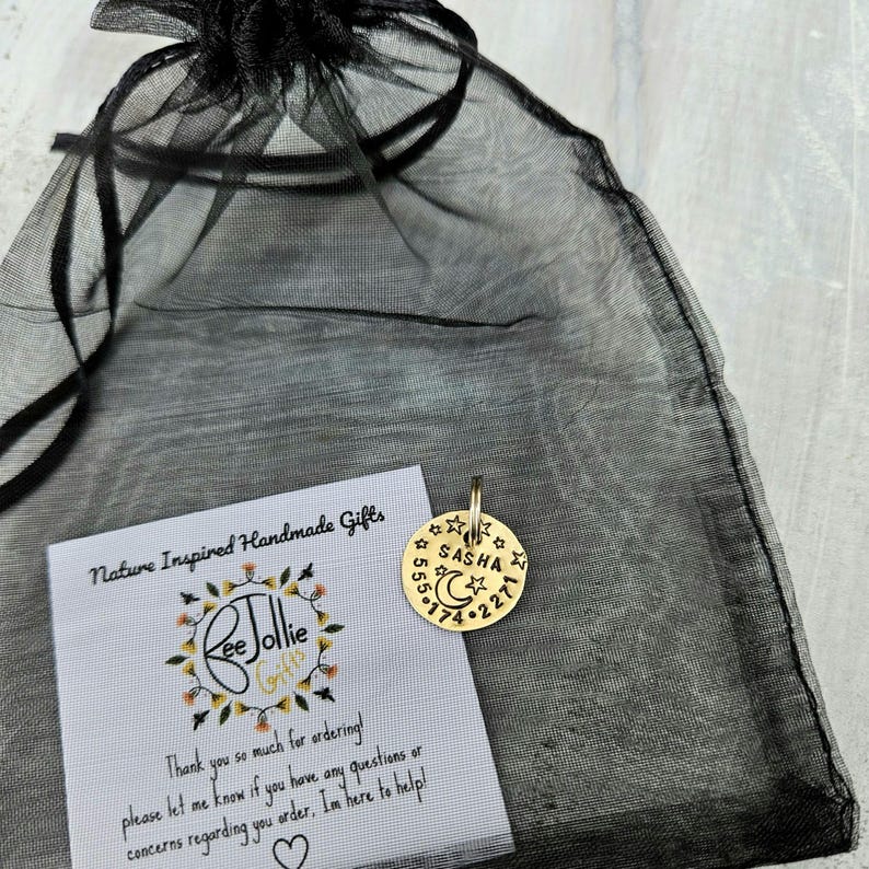May include: A round gold-toned metal pet tag featuring the name SASHA, a crescent moon, and stars, with the phone number 555-174-2271 engraved. The tag is placed on a black sheer organza drawstring bag next to a white card with the text ReeJollie Gifts.