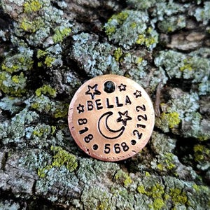 May include: A round copper pet ID tag featuring the stamped name BELLA, a crescent moon, and star icons. The tag includes the phone number 818-568-2122 engraved in a circular pattern around the centre design, with a small hole at the top for attachment.