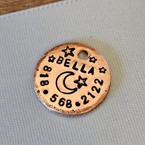 May include: A round copper-coloured metal pet identification tag featuring a hammered texture. The tag is engraved with the name BELLA, a crescent moon, three stars, and the phone number 818-568-2122. A small circular hole is located at the top.
