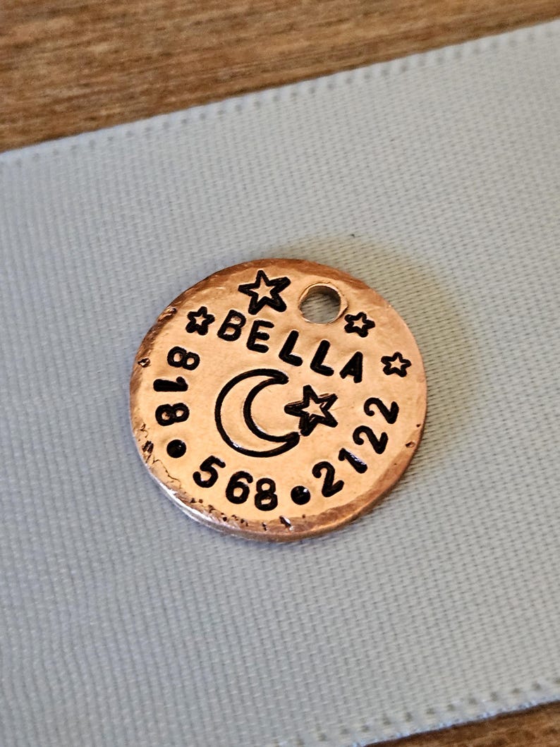 May include: A round copper-coloured metal pet identification tag featuring a hammered texture. The tag is engraved with the name BELLA, a crescent moon, three stars, and the phone number 818-568-2122. A small circular hole is located at the top.