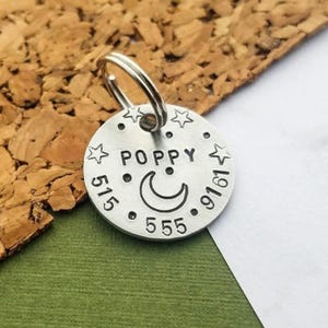 May include: A round silver-coloured metal pet ID tag featuring a split ring at the top. The surface is stamped with the name POPPY, a crescent moon, small stars, and the phone number 515-555-9161 in a clear, dark, sans-serif font style.