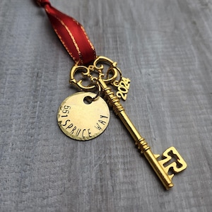 May include: A gold-colored key ornament with a red ribbon. A small gold tag is attached, inscribed with "551 SPRUCE WAY". A charm with "2024" is also attached. The key is likely a decorative item or a keepsake.