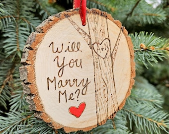 Op de afbeelding: Een houten ornament in de vorm van een boomstam met de tekst "Will You Marry Me?" en een hart met de initialen "A+N" erin gegraveerd. De ornament hangt aan een rood lint.