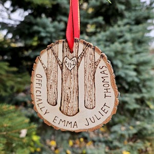 May include: A wooden ornament with a red ribbon, featuring three trees with a heart in the center. The text "Officially Emma Juliet Thomas 6-11-21" is engraved on the ornament.
