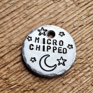 May include: A small, round, silver-colored metal tag with the words "MICRO CHIPPED" stamped in the center. The tag is decorated with stars and a crescent moon. A hole is at the top for attachment.