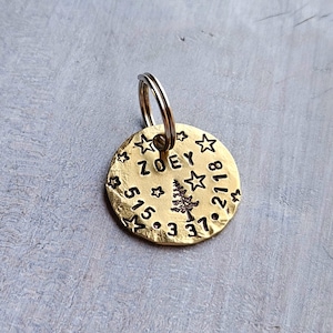 May include: A round, hammered brass pet ID tag featuring the name ZOEY, a pine tree graphic, and the numbers 515-337-2118 stamped into the surface. The tag is decorated with small star motifs and includes a silver-toned metal split ring.
