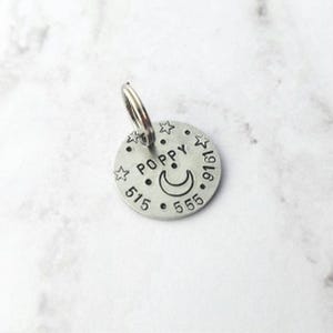 May include: A circular silver-toned metal pet identification tag featuring the stamped name POPPY, a crescent moon, and small stars. The tag includes the phone number 515-555-9161 and has a metal split ring attached to the top for easy collar attachment.