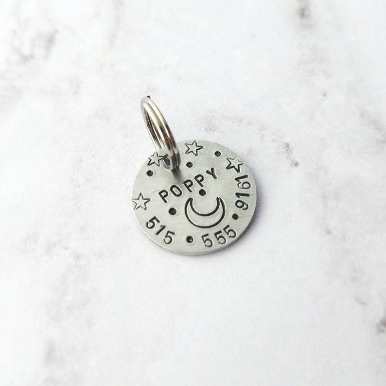 May include: A circular silver-toned metal pet identification tag featuring the stamped name POPPY, a crescent moon, and small stars. The tag includes the phone number 515-555-9161 and has a metal split ring attached to the top for easy collar attachment.