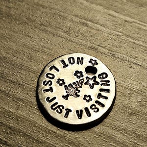 May include: A round silver-toned metal charm featuring the stamped text "NOT LOST JUST VISITING" in a circular pattern. The center includes a small stamped pine tree icon surrounded by three small star shapes, with a hole punched near the top edge.