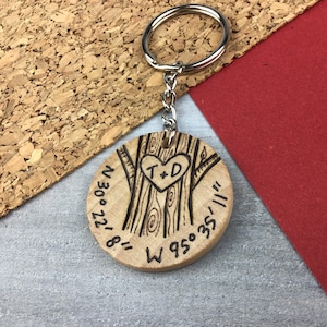May include: A wooden keychain with a silver ring and chain. The round wooden pendant features a tree design with a heart carved into the trunk, containing the initials "T+D". Coordinates and numbers are etched around the edge.