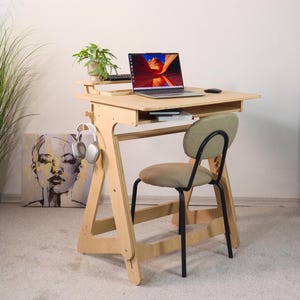 May include: A light-colored wooden standing desk with a laptop, keyboard, and small plant. A matching chair with a tan seat and black legs is positioned in front of the desk. White headphones hang on the side.