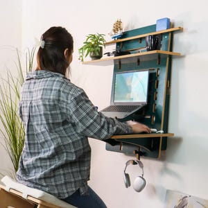 Puede incluir: Un escritorio montado en la pared de color verde oscuro con estantes, un ordenador portátil y una silla de rodillas de madera. El escritorio tiene varios estantes que sostienen una planta, una taza y otros objetos. Una persona está usando el portátil. Los auriculares cuelgan debajo.