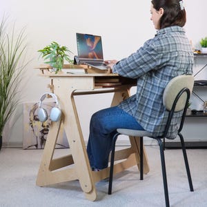 May include: A person sits at a wooden standing desk with a laptop, headphones, and a plant. The desk is made of plywood and has a simple design. The person is wearing a plaid shirt and jeans.