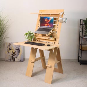 May include: A light-colored wooden standing desk with a laptop, keyboard, and mouse. The desk has multiple shelves and a headphone holder. A small plant sits on the desk. The desk is in a room with a painting and a tall plant.