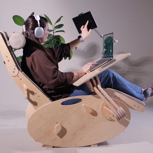 May include: A light-colored wooden rocking chair with an attached laptop desk and tablet holder. The chair has a headrest and is designed for comfort. A person is using the chair with a laptop and tablet. The chair is made of light-colored wood.