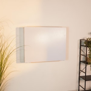 May include: A rectangular, white, wall-mounted folding table in a closed position. The table features a smooth, solid white surface and is mounted flush against a plain wall, appearing as a flat panel when folded down to save space.