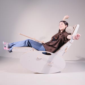 Peut inclure: Un fauteuil à bascule en bois clair avec une base incurvée et un dossier réglable. La chaise a un appui-tête et un repose-pieds. La personne sur l'image porte un jean bleu et une chemise marron.