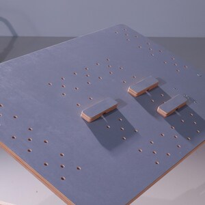 May include: A gray wooden board with numerous small holes and three rectangular wooden blocks. The edges of the board are a natural wood color. The board is likely a component of a larger structure or a specialized work surface.