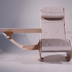 K&ouml;nnte beinhalten: Ein hellfarbiger Holzstuhl mit einem angebrachten Schreibtisch. Der Stuhl hat eine gepolsterte R&uuml;ckenlehne und einen Sitz aus neutralem Stoff. Der Schreibtisch ist rechteckig mit kleinen L&ouml;chern und wird von einem gebogenen Holzarm getragen.