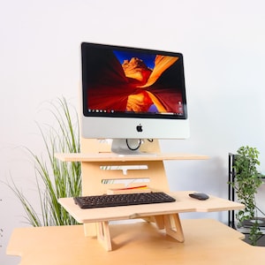 Może przedstawiać: Drewniane biurko stojące z białym komputerem iMac na górze. Biurko ma dwie półki, a klawiatura i mysz znajdują się na dolnej półce. Biurko stoi na jasnym drewnianym blacie.