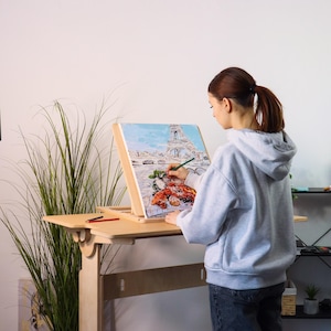 May include: A person paints on a canvas with a wooden easel. The canvas depicts a painting of the Eiffel Tower in Paris, France. The easel is attached to a wooden standing desk.