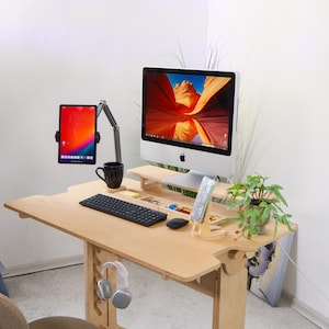 May include: A light wood standing desk with a computer monitor, a tablet, a keyboard, a mouse, a phone stand, a cup, and headphones. The desk has a built-in shelf and a drawer.
