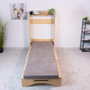 Peut inclure: Un lit pliable en bois avec un matelas gris. Le lit est équipé d'une étagère en haut avec une tasse noire, un petit panier et une plante. Le lit est conçu pour gagner de la place.