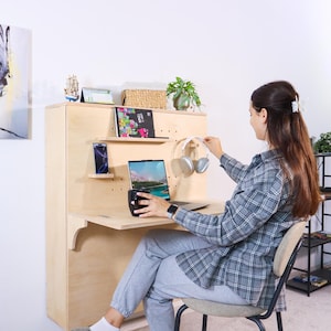 May include: A light-colored wooden wall-mounted desk with shelves, a laptop, and a smartphone. A person is holding a pair of white headphones. A black mug sits on the desk. A small plant and a decorative sailboat are on top of the desk.