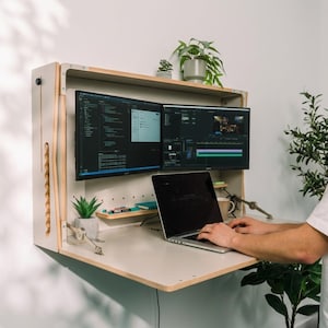 May include: A wall-mounted, fold-down wooden desk featuring a light-colored finish, two mounted computer monitors displaying software interfaces, a laptop on the work surface, a small potted plant on an upper shelf, and metal support hinges.