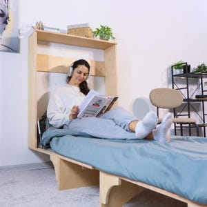 Op de afbeelding: Een lichtgekleurd, houten wandbed met een blauwe matras. Het bed is uitgeklapt en een persoon zit erop en leest een tijdschrift. Een plank boven het bed bevat een mand, planten en andere items. De persoon draagt een koptelefoon.