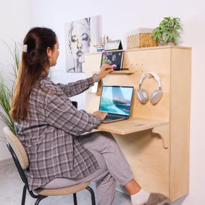 May include: A light-coloured wooden wall-mounted desk with a laptop, tablet, and headphones. The desk has a small shelf and a fold-down desk surface. A person is sitting at the desk, working on the laptop. A painting and a plant are visible.