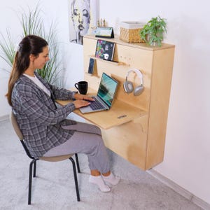 May include: A wall-mounted wooden desk with a fold-down work surface. The desk has shelves for a tablet and phone, and a pair of headphones hangs on the side. A laptop sits on the desk, and a person is seated in front of it.