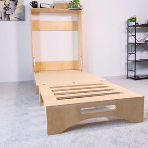 May include: A light-coloured, wooden wall bed in the open position. The bed frame is rectangular with a slatted footboard. The headboard section is attached to the wall and has a shelf with decorative items.