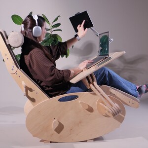 K&ouml;nnte beinhalten: Ein hellfarbener Schaukelstuhl aus Holz mit einem angebrachten Laptop-Schreibtisch und Tablethalter. Der Stuhl hat eine Kopfst&uuml;tze und verstellbare Funktionen. Eine Person benutzt den Laptop und das Tablet, w&auml;hrend sie Kopfh&ouml;rer tr&auml;gt. Eine Pflanze ist im Hintergrund.