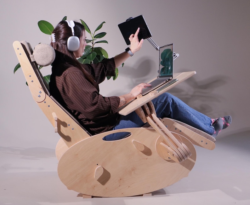 K&ouml;nnte beinhalten: Ein hellfarbener Schaukelstuhl aus Holz mit einem angebrachten Laptop-Schreibtisch und Tablethalter. Der Stuhl hat eine Kopfst&uuml;tze und verstellbare Funktionen. Eine Person benutzt den Laptop und das Tablet, w&auml;hrend sie Kopfh&ouml;rer tr&auml;gt. Eine Pflanze ist im Hintergrund.