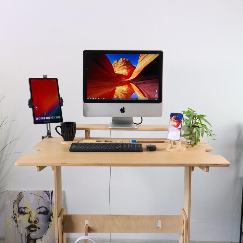 May include: A light wood adjustable standing desk with a computer, tablet, and smartphone. The computer screen displays a landscape image. A black mug, keyboard, and mouse are on the desk. A small plant and artwork are also present.