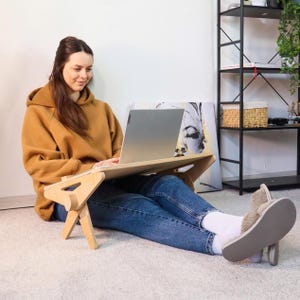 以下が含まれることがあります： 折りたたみ式の脚が付いた、明るい色の木製ラップトップデスク。 ベッドや床での使用を想定して設計されています。 ラップトップはシルバーで開いています。 茶色のパーカーとブルージーンズを着ています。