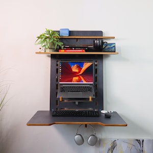 May include: A black wall-mounted desk with a laptop, keyboard, and headphones. The desk has two shelves, one above the other, with a plant, books, and a mug on the top shelf.