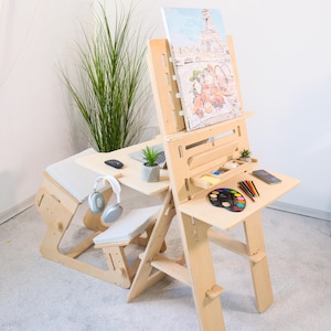 May include: A light-colored wooden artist's desk and easel combination. The desk has a laptop, mouse, headphones, and a small plant. The easel holds a painting of the Eiffel Tower. A palette of paints and colored pencils are on the side.