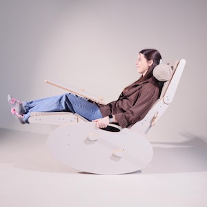 May include: A light-colored wooden rocking chair with an attached desk. The chair has a curved base, a padded headrest, and adjustable armrests. The person in the chair is wearing a brown jacket and blue jeans.